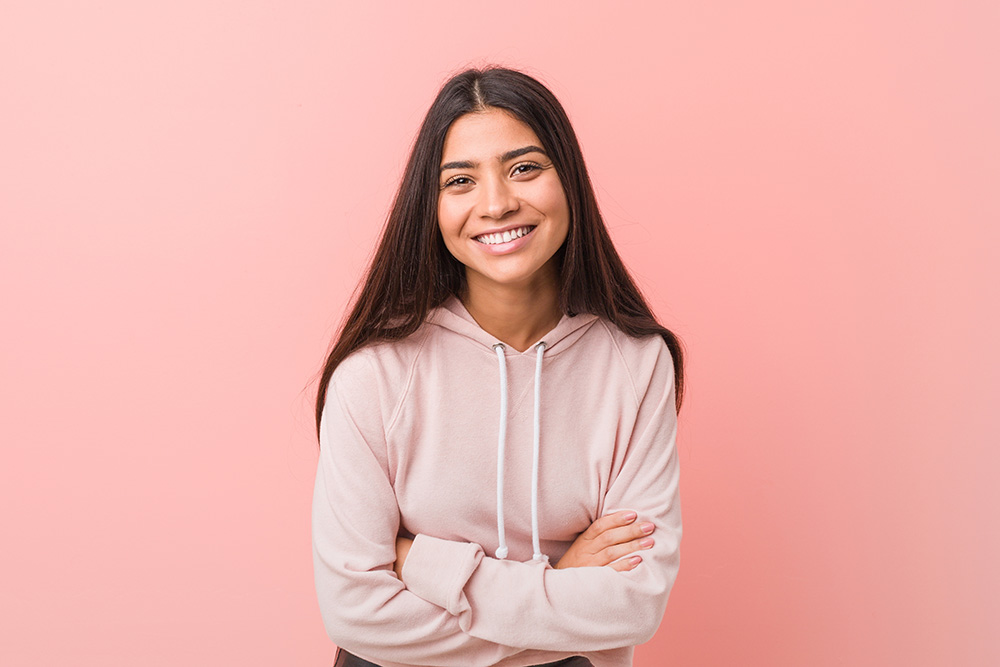 A woman with a smile stands against a pink background, wearing a light-colored hoodie with her arms crossed.